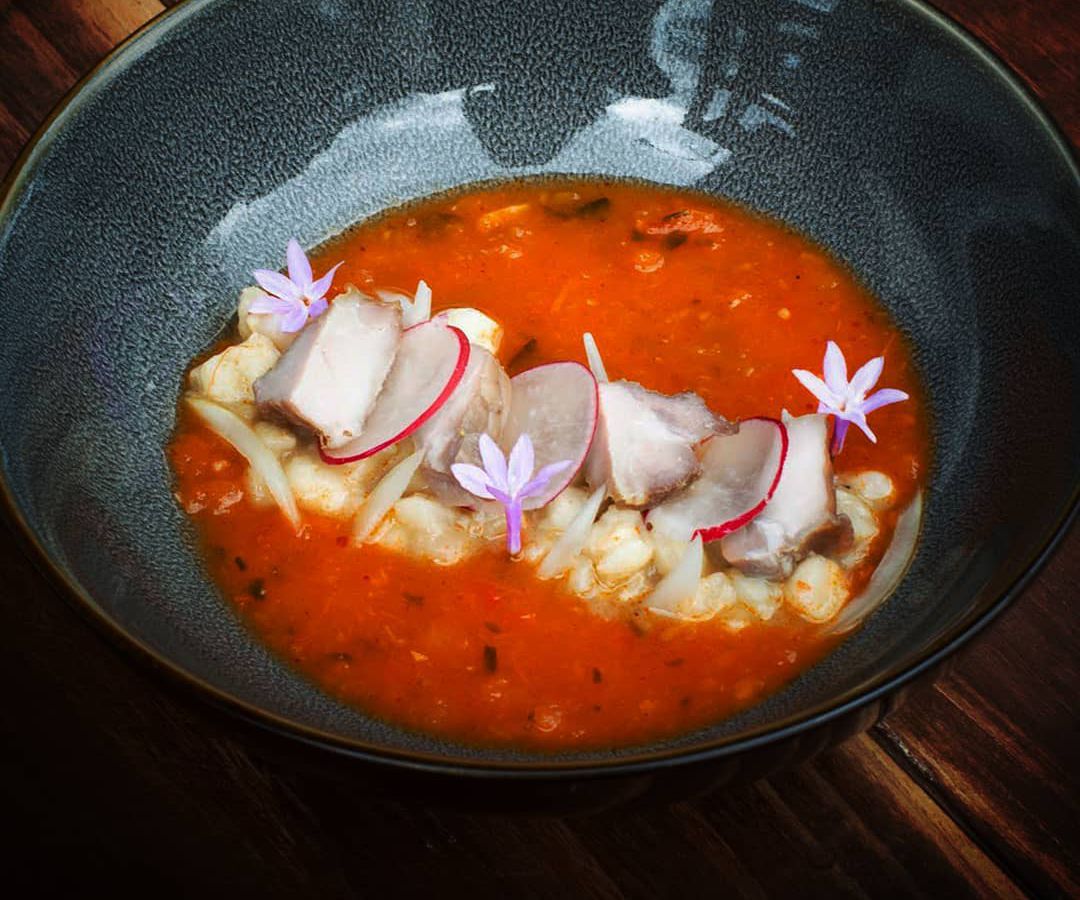 A bowl of bright broth with slices of pork, radish, and hominy