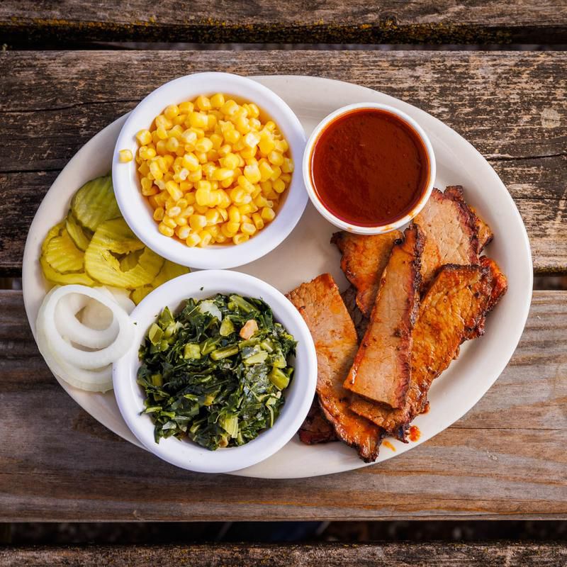 A plate holds, clockwise: a dish with corn, a dish with barbecue sauce, barbecue chicken, greens, and raw onions and pickles.