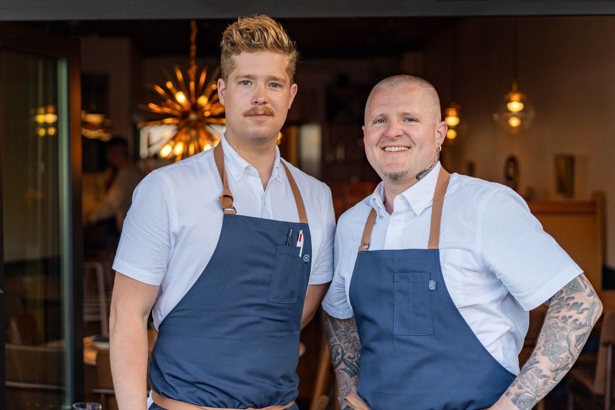 Two men stand next to each other wearing light blue shirts and dark blue aprons.
