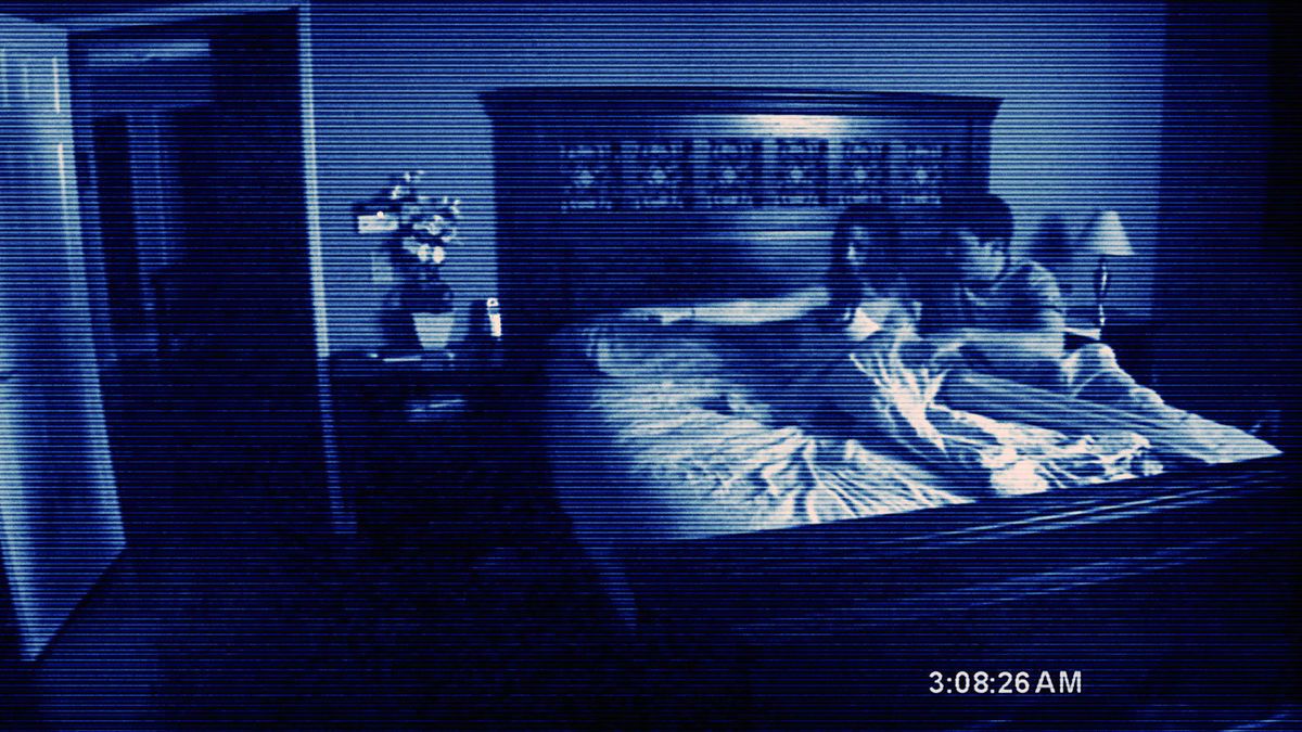 A couple huddles in bed, pointing fearfully at something offscreen, in a grainy, low-resolution camera image from 2007’s Paranormal Activity