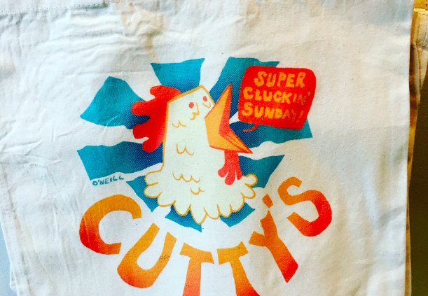 Closeup on a white tote bag with blue and orange art, featuring a chicken yelling “super cluckin’ sunday”