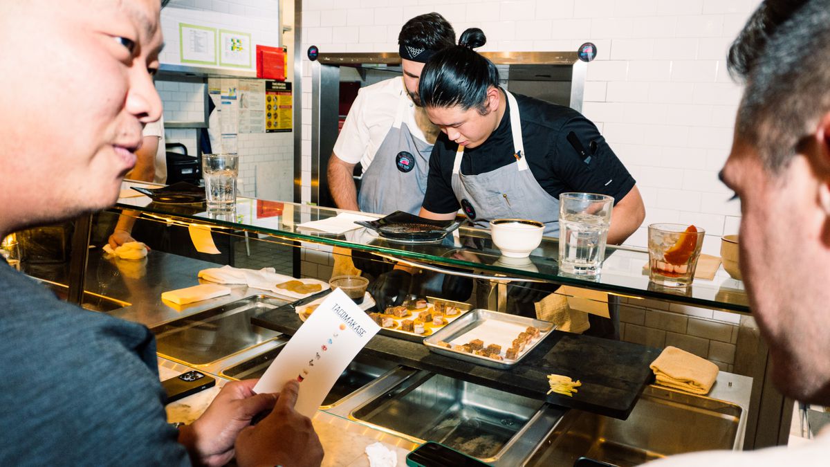 Customers sit behind a sneeze guard as two chefs prepare tacos.