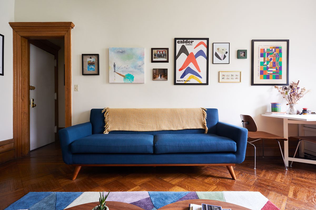 A living room with a blue midcentury-style couch in the center. A colorful geometric rug sits afoot, above herringbone floors. Various artworks hang on white walls.
