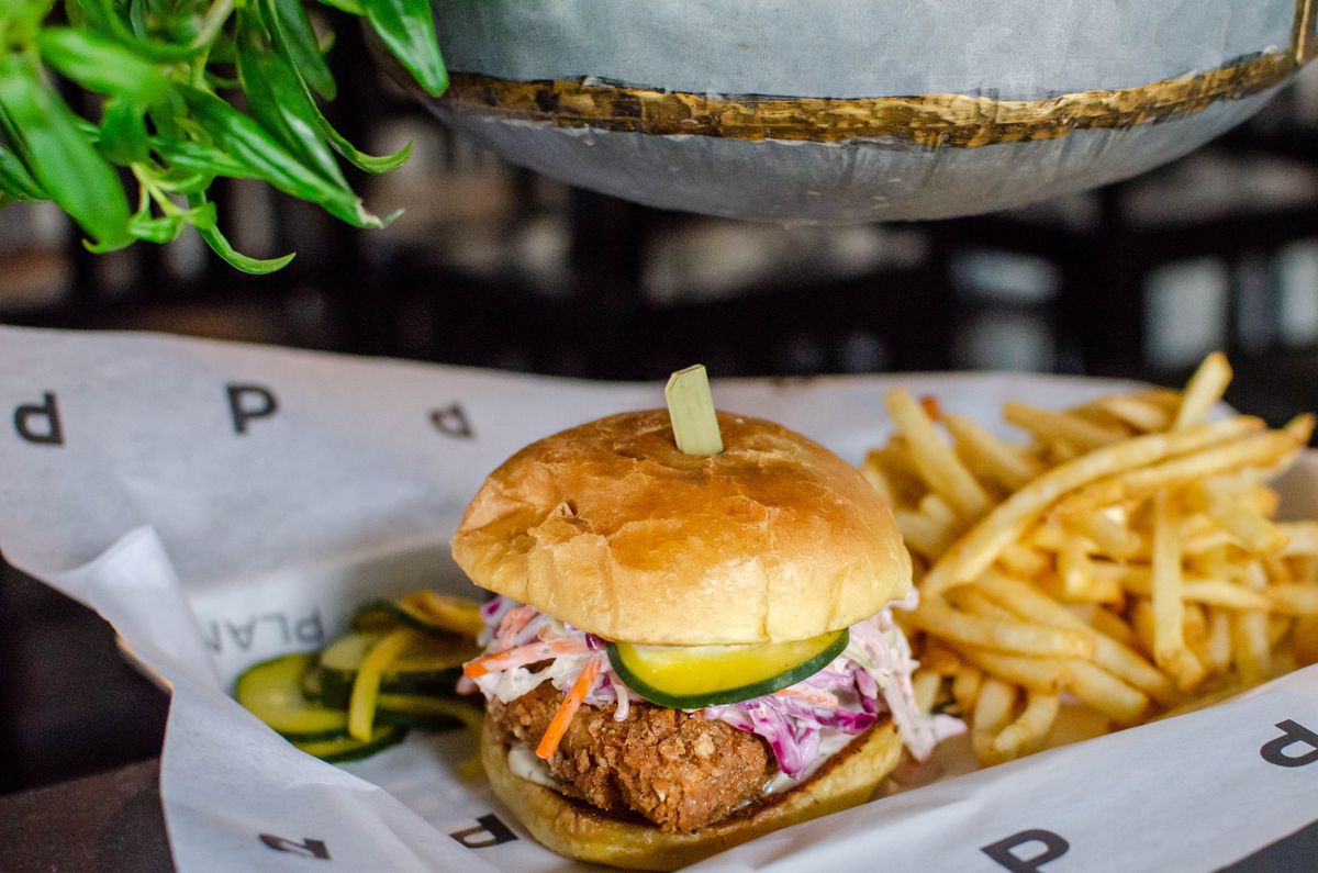 A patty of vegan fried “chicken” with a side of fries sits on white paper on a silver tray.