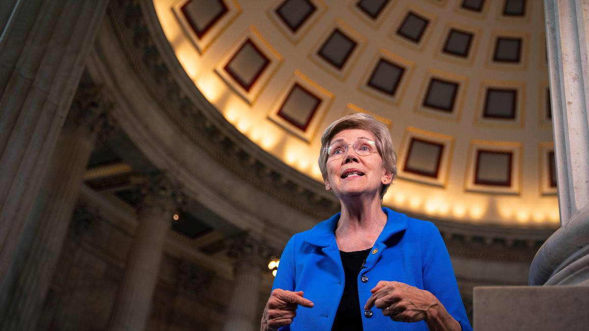 Sen. Elizabeth Warren is shown speaking in the Capitol rotunda to reporters, wearing a blue jacket and framed by the ceiling and pillars.