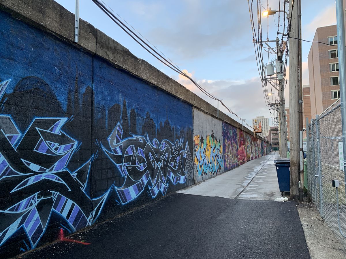 Looking north in the alley towards West Sheridan Road, a mural by Eric Villarreal, among other artists, shows a dark cityscape background and stylized lettering.