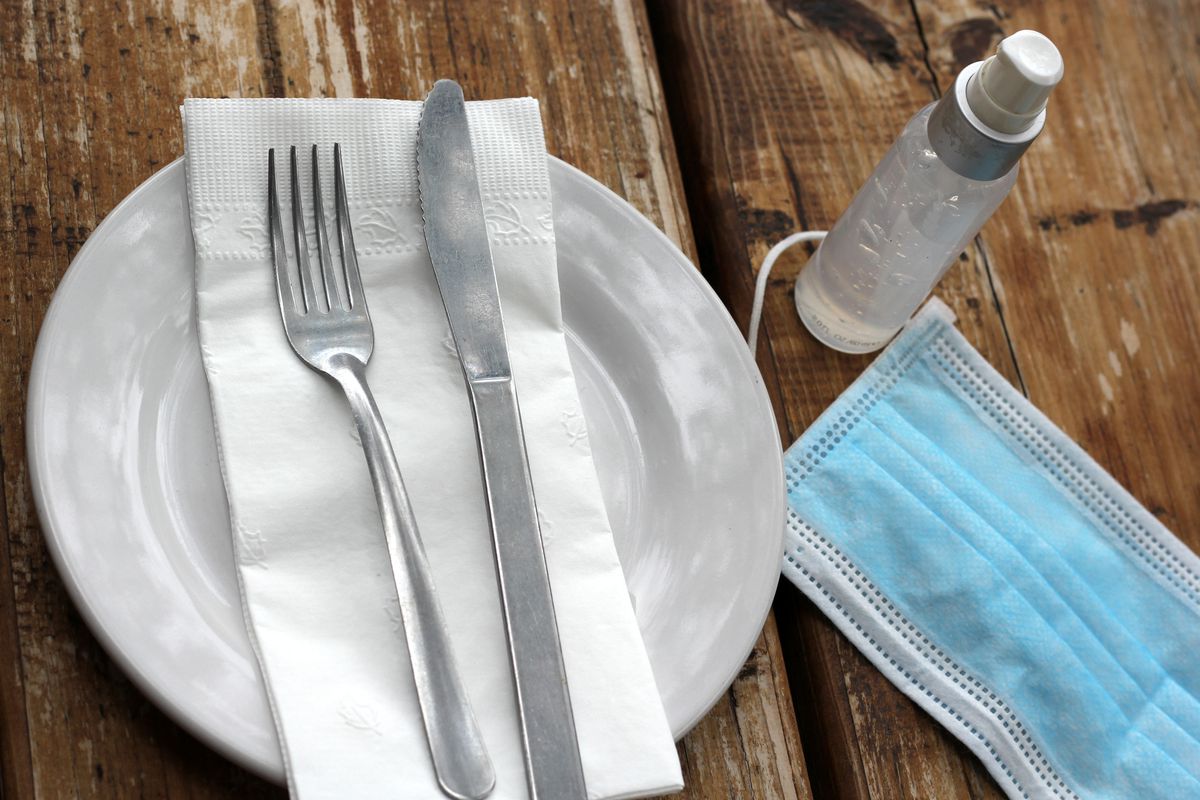 A plate with a fork, knife, and napkin on top, next to a bottle of hand sanitizer and a light blue surgical mask