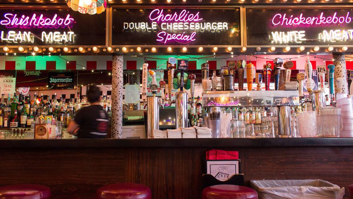 A restaurant interior features red leather stools at a bar. Neon signage over a mirror in the background advertises Charlie’s double cheeseburger special.