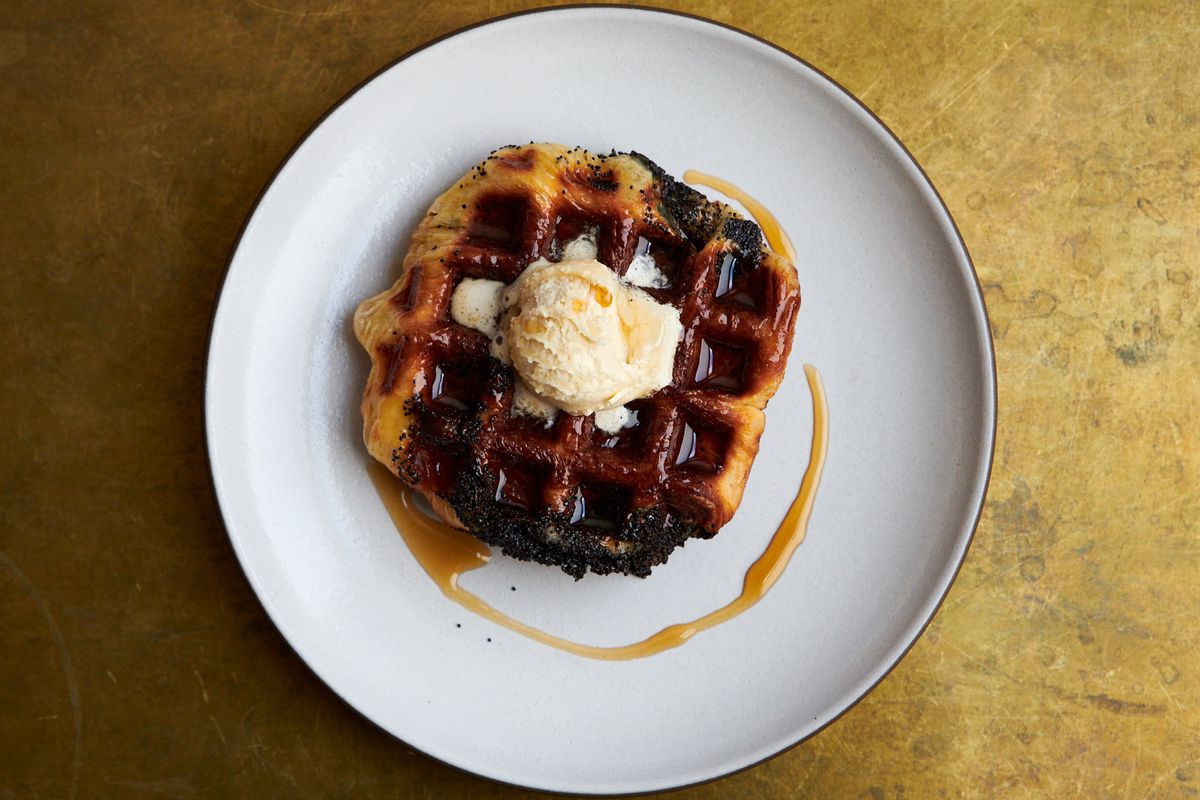 A waffle with a dollop of maple butter and swirl of maple syrup sits on a white plate. It’s browned with caramelization and somewhat misshapen.