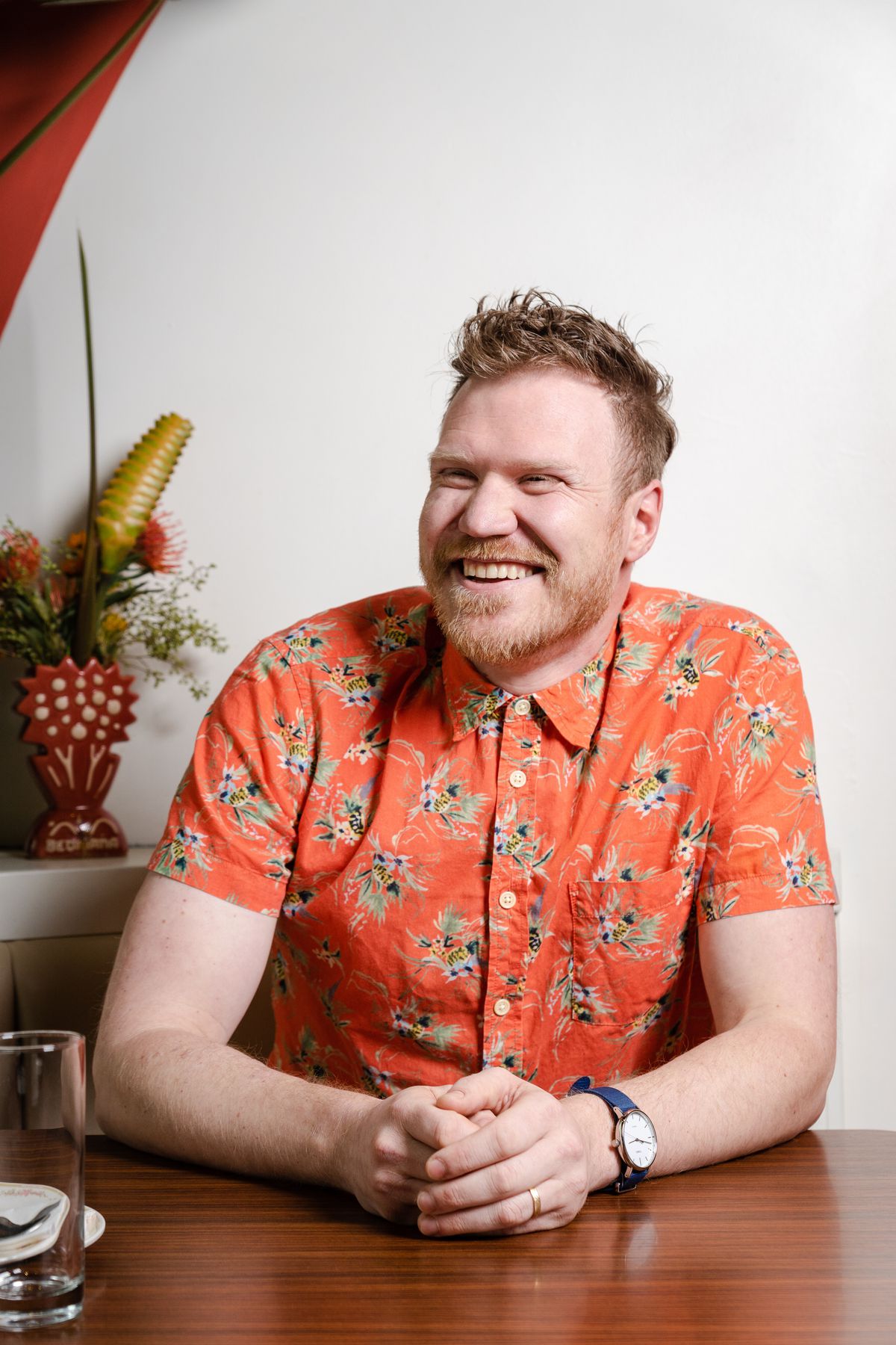 A man sits, laughing, at a table with his hands folded, wearing a blue watch and a bright orange button up with short sleeves. He’s smiling, with a short strawberry blonde beard