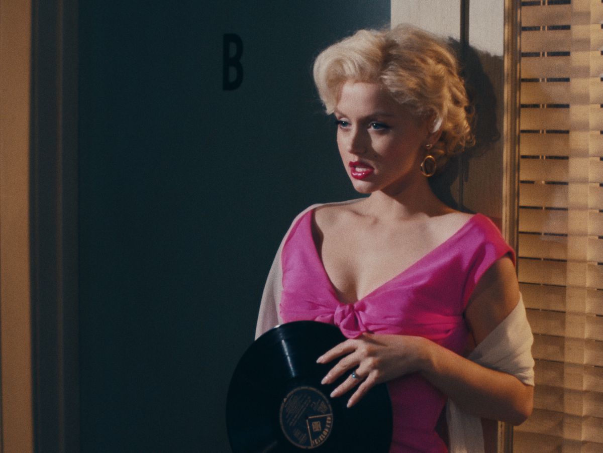 A woman with platinum blonde hair, dressed in a pink satin gown, leans against a doorway. Her eyes are half-closed, her mouth is half-open, and she is holding a record.