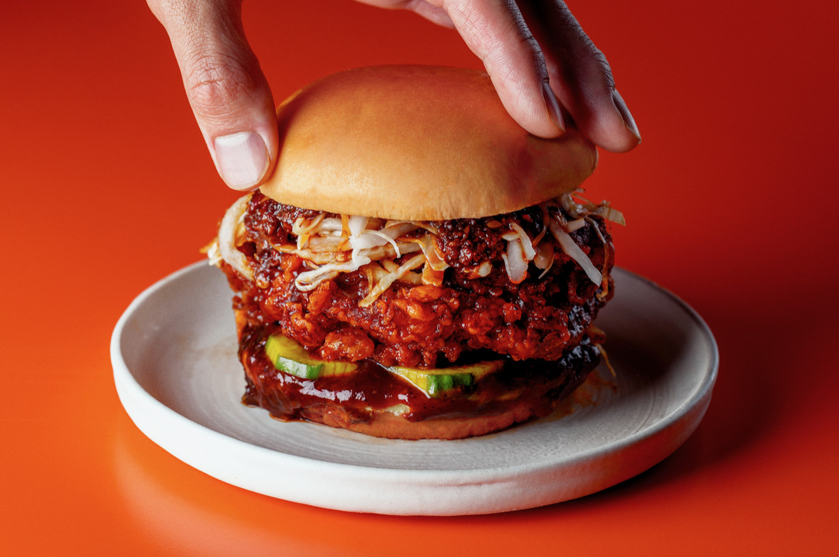 A hand presses the top bun down on a fried chicken sandwich glistening with chilli oil and doubanjiang.