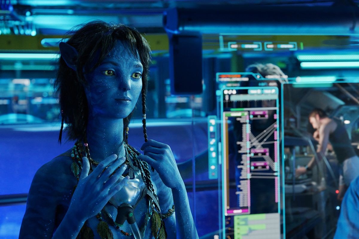 Kiri holds her braid as she stands in a human laboratory watching a video from her “mom” Dr. Grace Augustine (Sigourney Weaver) speaking on a computer screen in Avatar: The Way of Water
