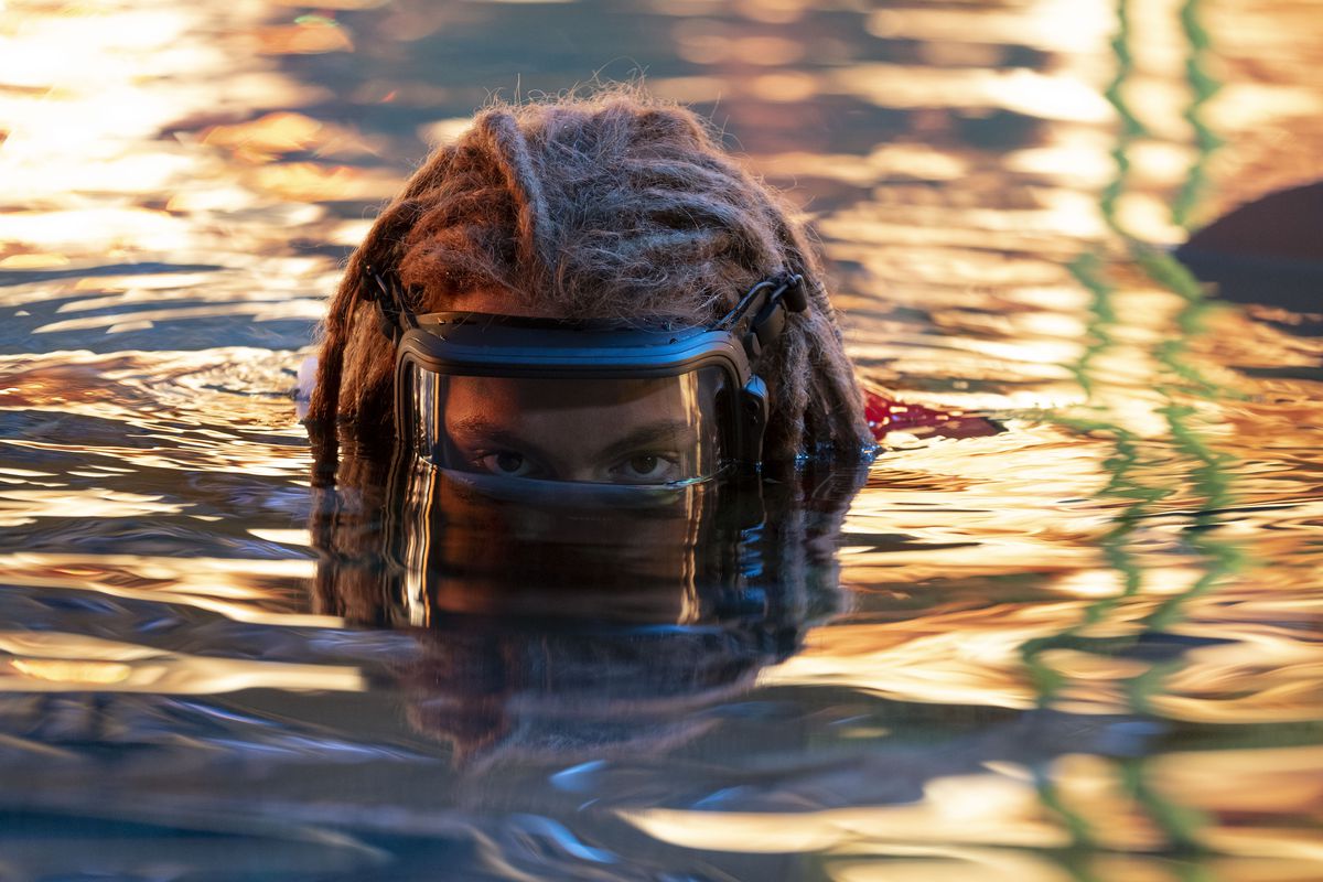 Spider (Jack Champion) wearing an oxygen mask as he rises out of the water, which shimmers with fire reflections in Avatar: The Way of Water