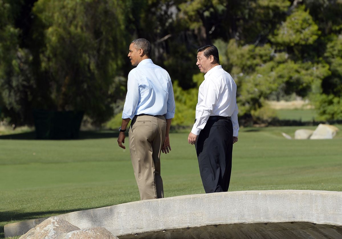 Obama and Xi walk along a smooth grass lawn with trees and landscaped rocks in the background. Obama wears khaki pants and a blue collared shirt with the sleeves partly rolled up, while Xi wears navy slacks and a white collared shirt. Both are jacketless and tieless.