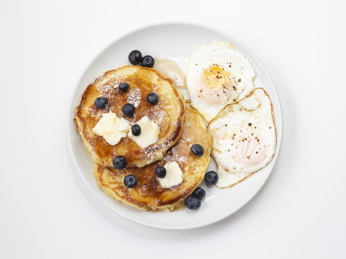 A plate with two blueberry pancakes garnished with blueberries and butter and two fried eggs