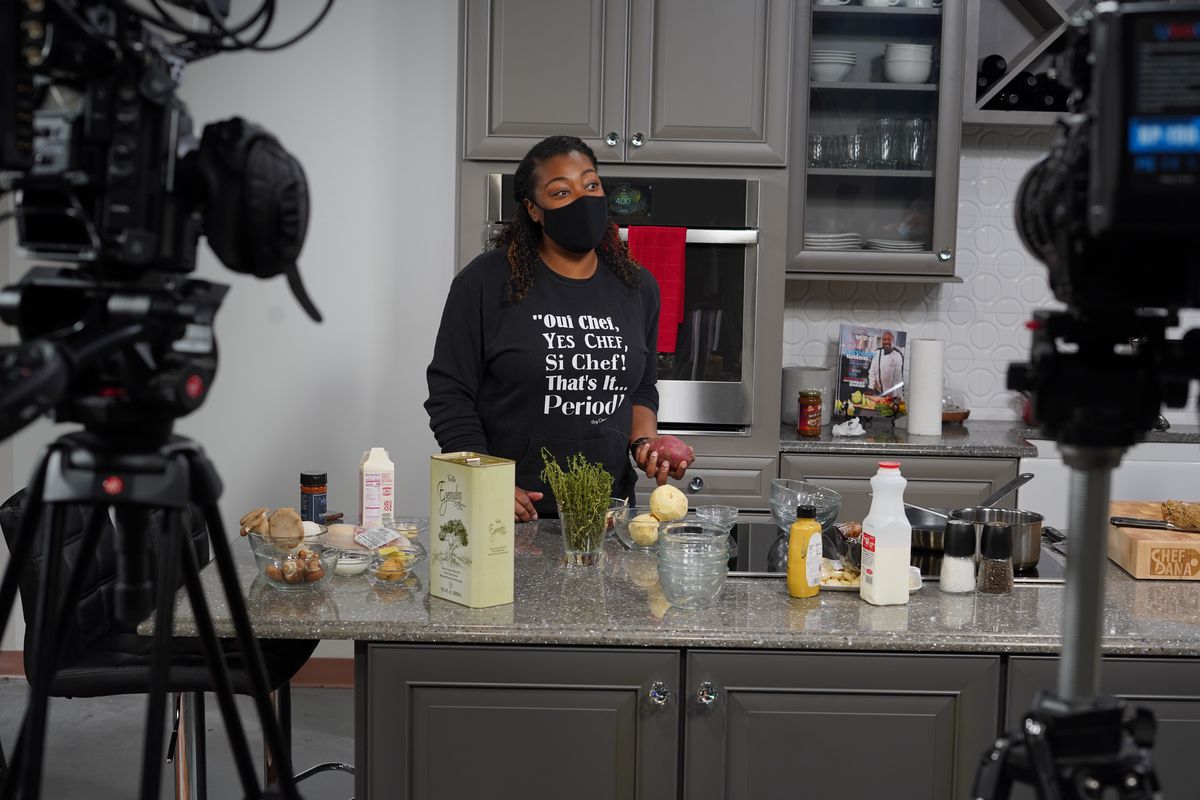 A Black woman wearing a black shirt and black face mask holds a potato while standing in a kitchen. Ingredients like olive oil are scattered on the counter, and a pair of cameras sits in the background.