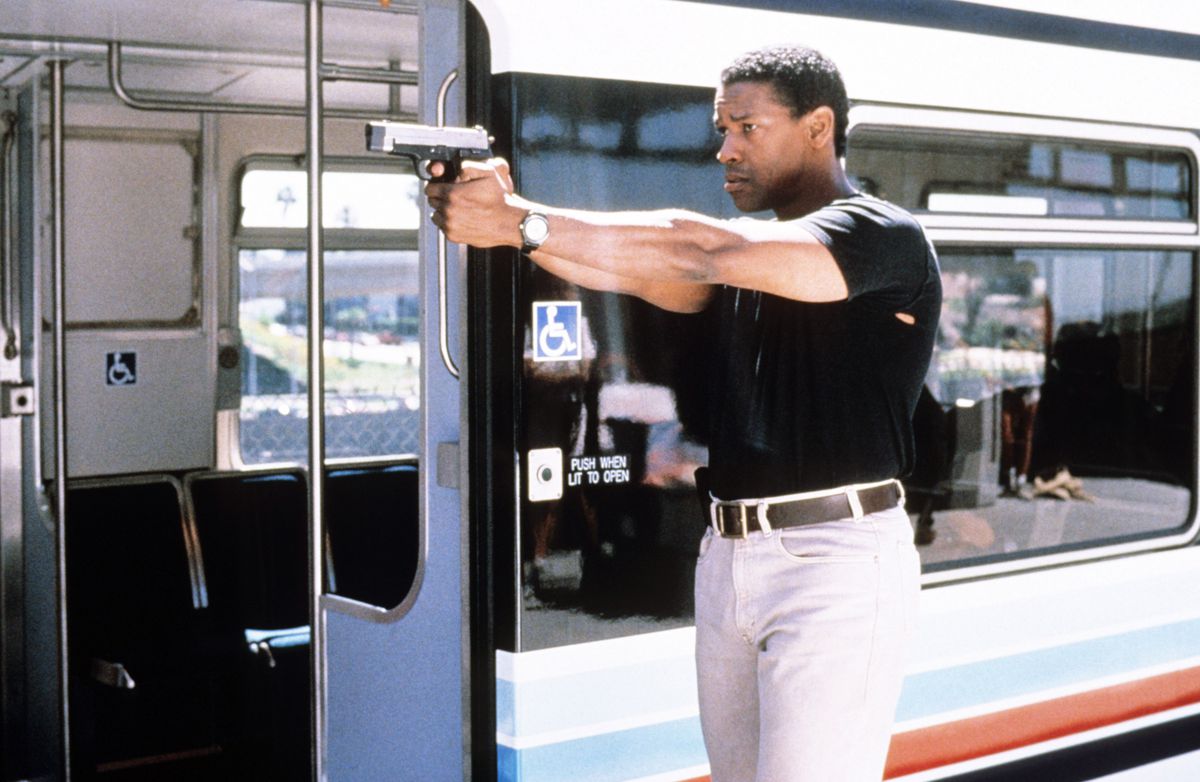 Denzel Washington, wearing a tight black t-shirt, holds a gun next to a train car in Virtuosity.