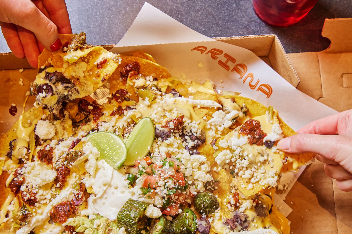 A cardboard box filled with nachos with two hands grabbing chips from either side of the box.