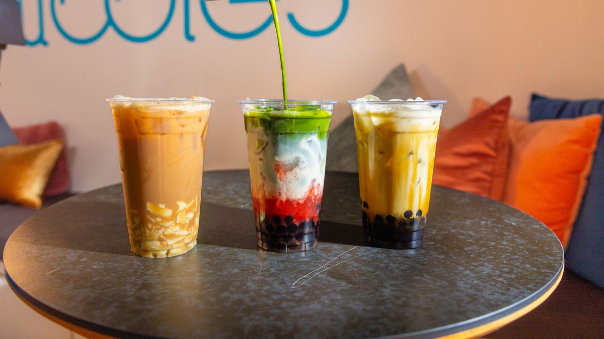 Three multi-color boba tea plastic cups lined up in a horizontal row on a gray table set against a white wall with a blue Eat Bubbles logo