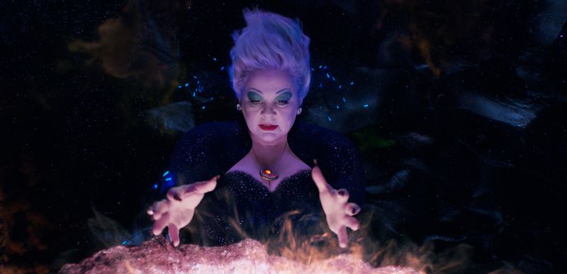 Melissa McCarthy as Ursula. She’s lit in purple light and her hands work over a smoking cauldron.