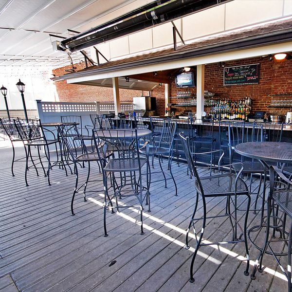 A restaurant’s covered roof deck has black metal tables and chairs, a brick wall, and a full bar.