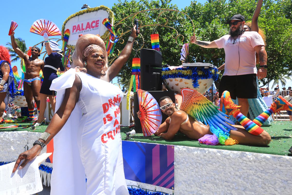 Drag personality Tiffany Fantasia stands in front of a colorful parade float dressed in a white gown featuring the words “Drag is not a crime” across the chest in red fabric.