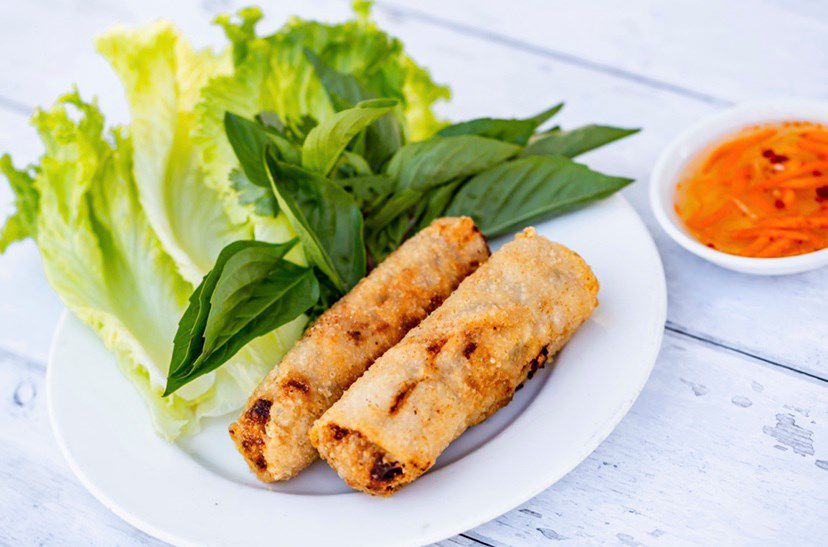 Two spring rolls with lots of herbs on the plate and nuoc cham dipping sauce to the right.