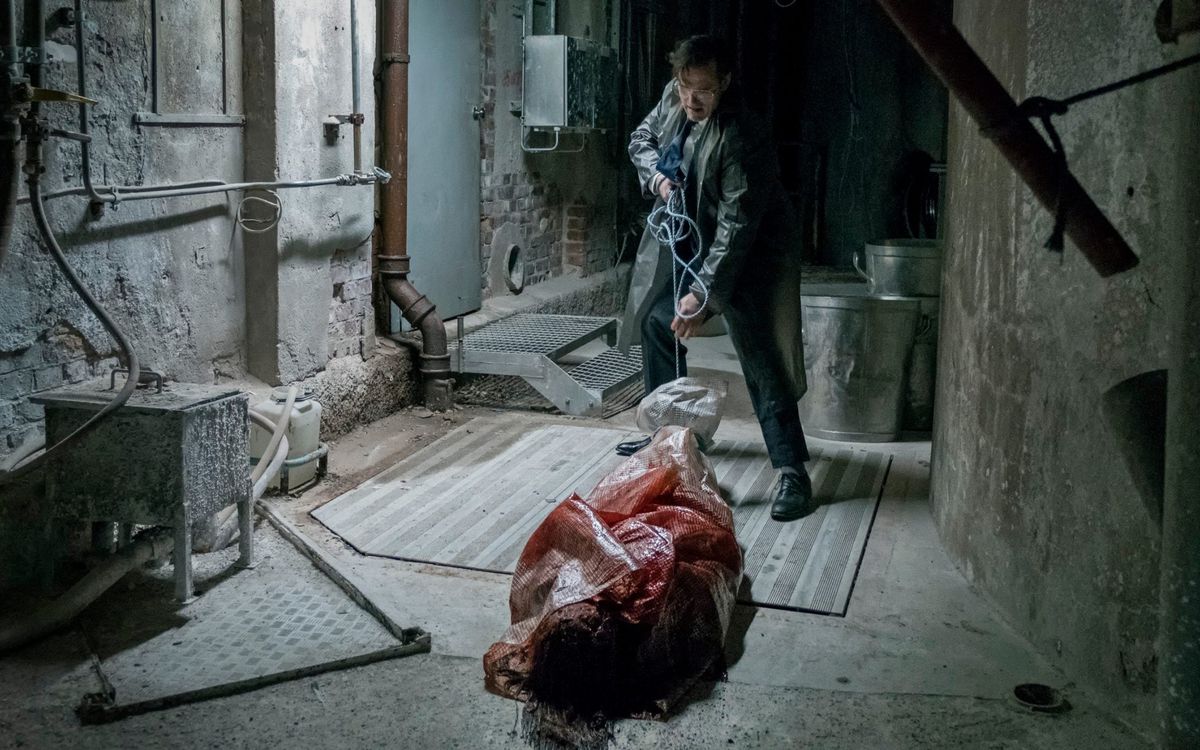 Matt Fillon, in a suit and plastic raincoat, drags a plastic-wrapped, blood-soaked corpse through a dirty basement space in The House that Jack Built.
