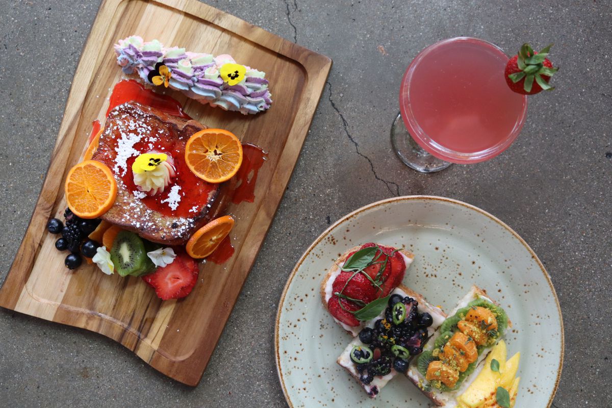 A wooden board with Fruity Pebble french toast, a plate of tropical fruit toast, and a Loud &  Proud cocktail with a strawberry garnish.
