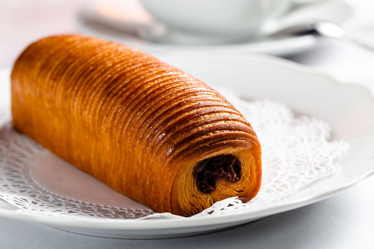 A golden, cylinder-shaped chocolate croissant is photographed diagonally, highlighting the ribbed texture of the pastry and its chocolate interior, visible from the front end