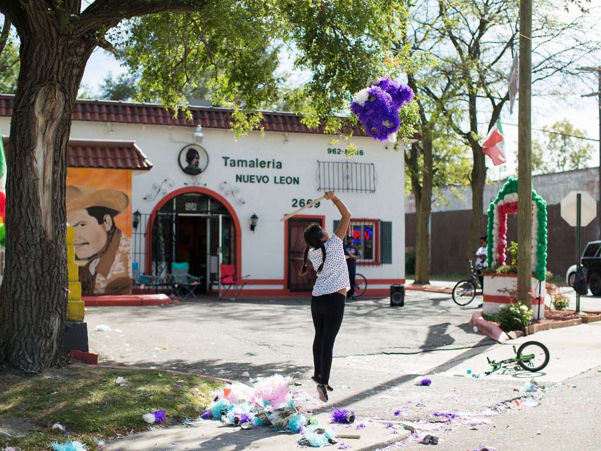 A girl in a white shirt and black jeans is shown mid-air jumping to hit a purple piñata in front of the white and red Tamaleria Nuevo Leon restaurant.
