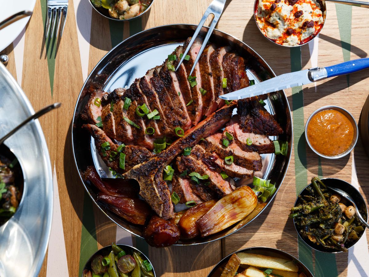 A sliced T-bone steak arranged in a circular metal platter with cups of salatim and dips placed around the platter.