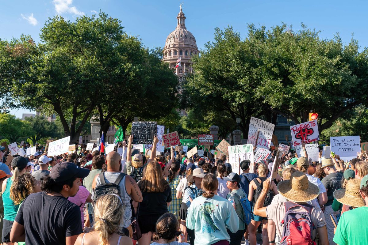 The Greco-Roman Capitol dome peeks through green trees under a blue sky; the US and Texas flags fly over it. In the foreground of the photo, a dense knot of protesters (seen from behind) appear to be marching towards the Capitol.