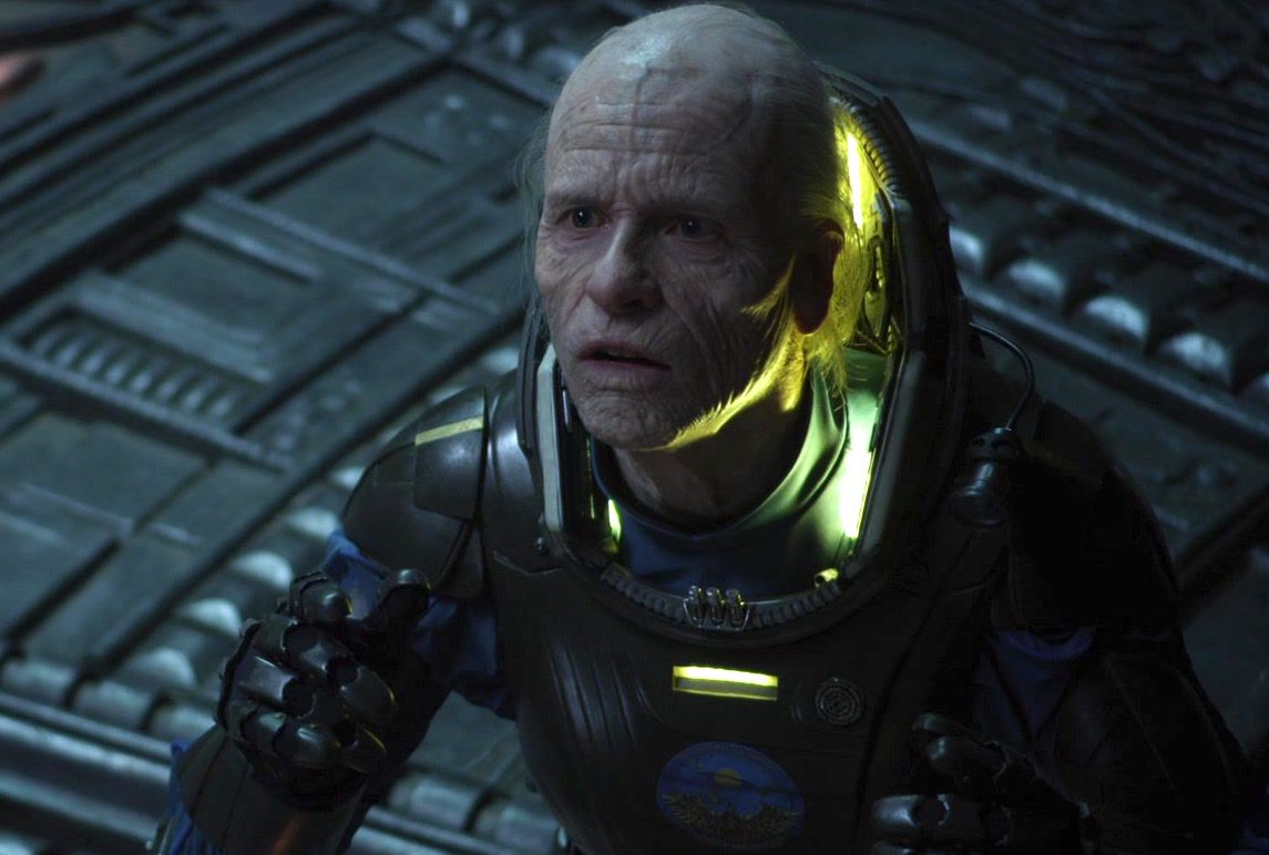Old Weyland (Guy Pearce con maquillaje de vejez) con un traje espacial inclinándose ante una presencia fuera de la pantalla en una nave espacial extraterrestre en Prometheus