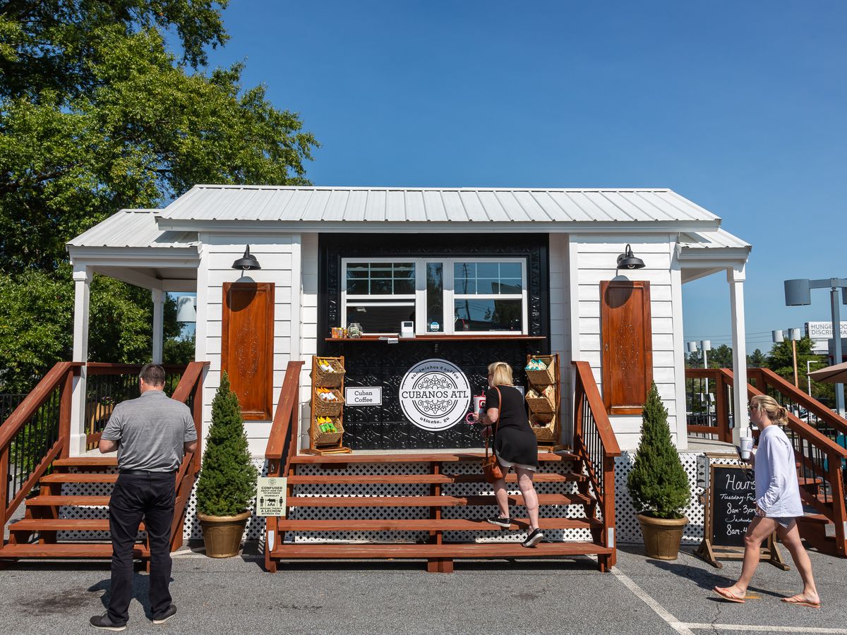 A white tiny house with two to-go windows framed in black paint with people standing in front of it masked waiting to get their takeout orders