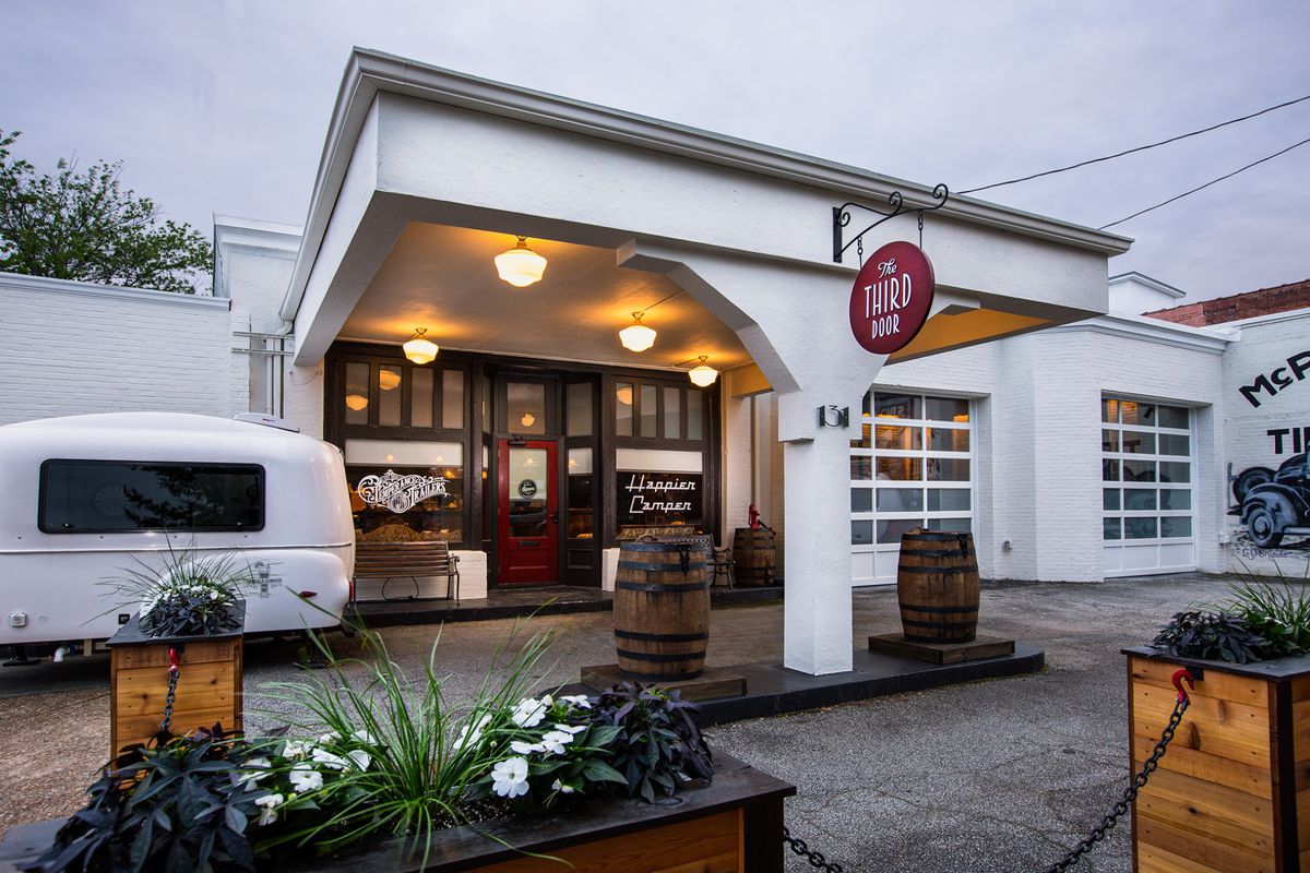 A white retro camper beside a bright white portico with large dark wood double doors at a bar in Marietta, GA, once an old gas station with two bourbon barrels where the gas pumps would be and a red sign hanging overhead with the name The Third Door