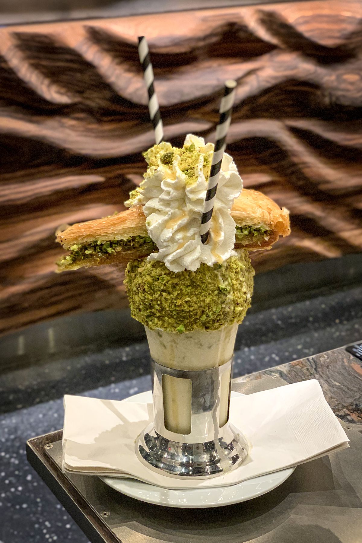 A vertical photo of a shake with whipped cream, two straws, and a triangle of baklava on top.