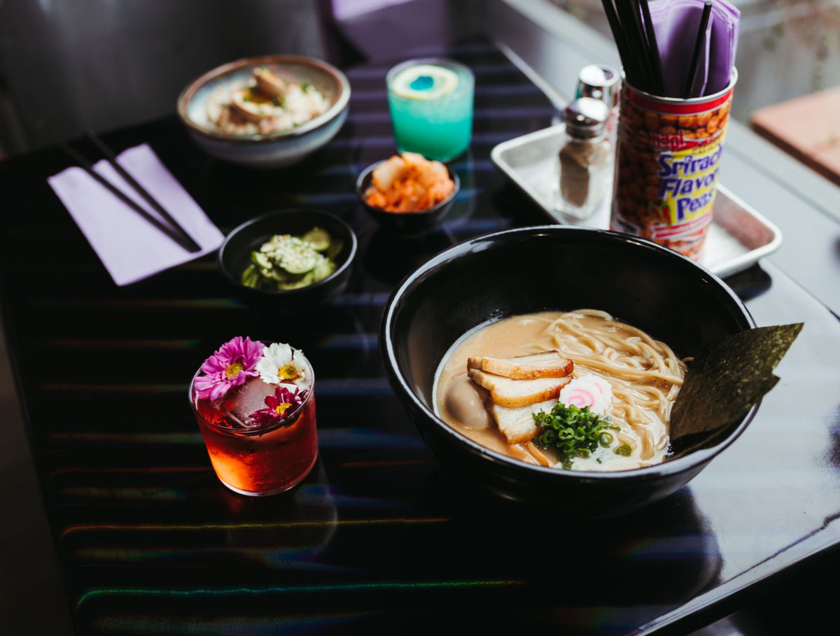 A table lined with ramen, potato salad, cucumber salad, kimchi, and two cocktails.