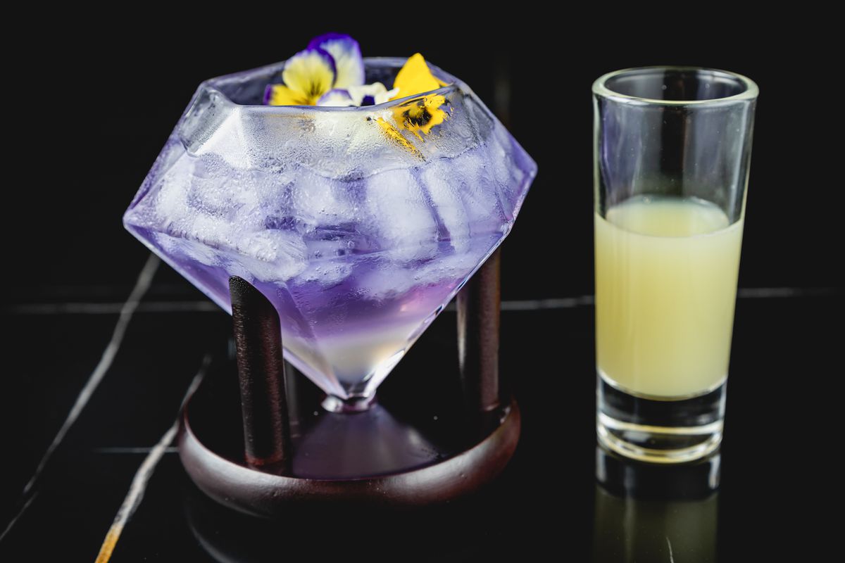 MaKiin's Karne Blue cocktail is served in a diamond-shaped glass topped with flowers and served with a shot on the side.