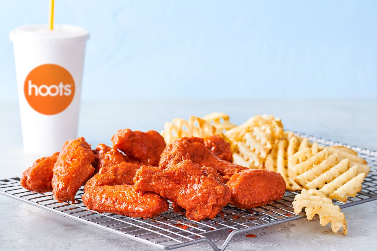A metal rack holds buffalo wings and waffle fries beside a white paper soda cup that reads “Hoots.”