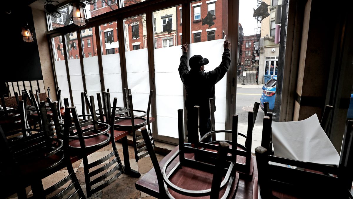 Man putting up paper over restaurant windows amid chairs stacked on top of tables.