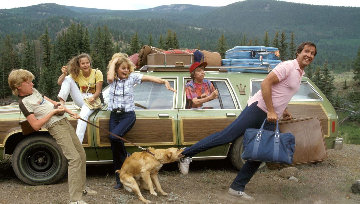 The Vacation cast poses in front of their ugly green station wagon, with Chevy Chase holding up a couple of suitcases as the family dog they’re transporting pulls on his pant leg with its teeth.