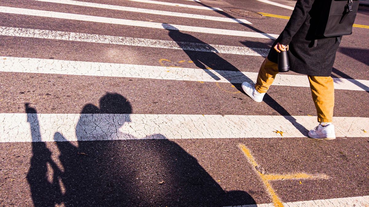 A low-to-the-ground angle shows the torso and legs of a person walking across the street on a crosswalk, and the silhouette shadow of a person in a wheelchair.