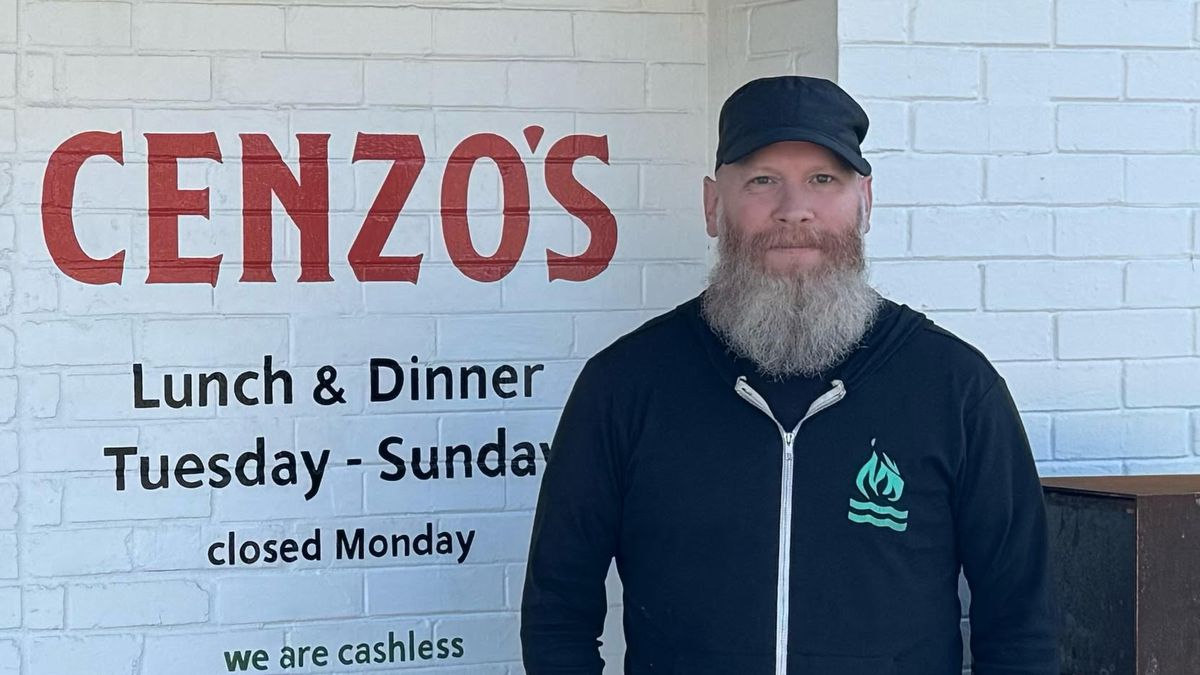 A man with a beard stands in front of a building. “Cenzo’s” is painted on the brick building, along with its hours of operation. 