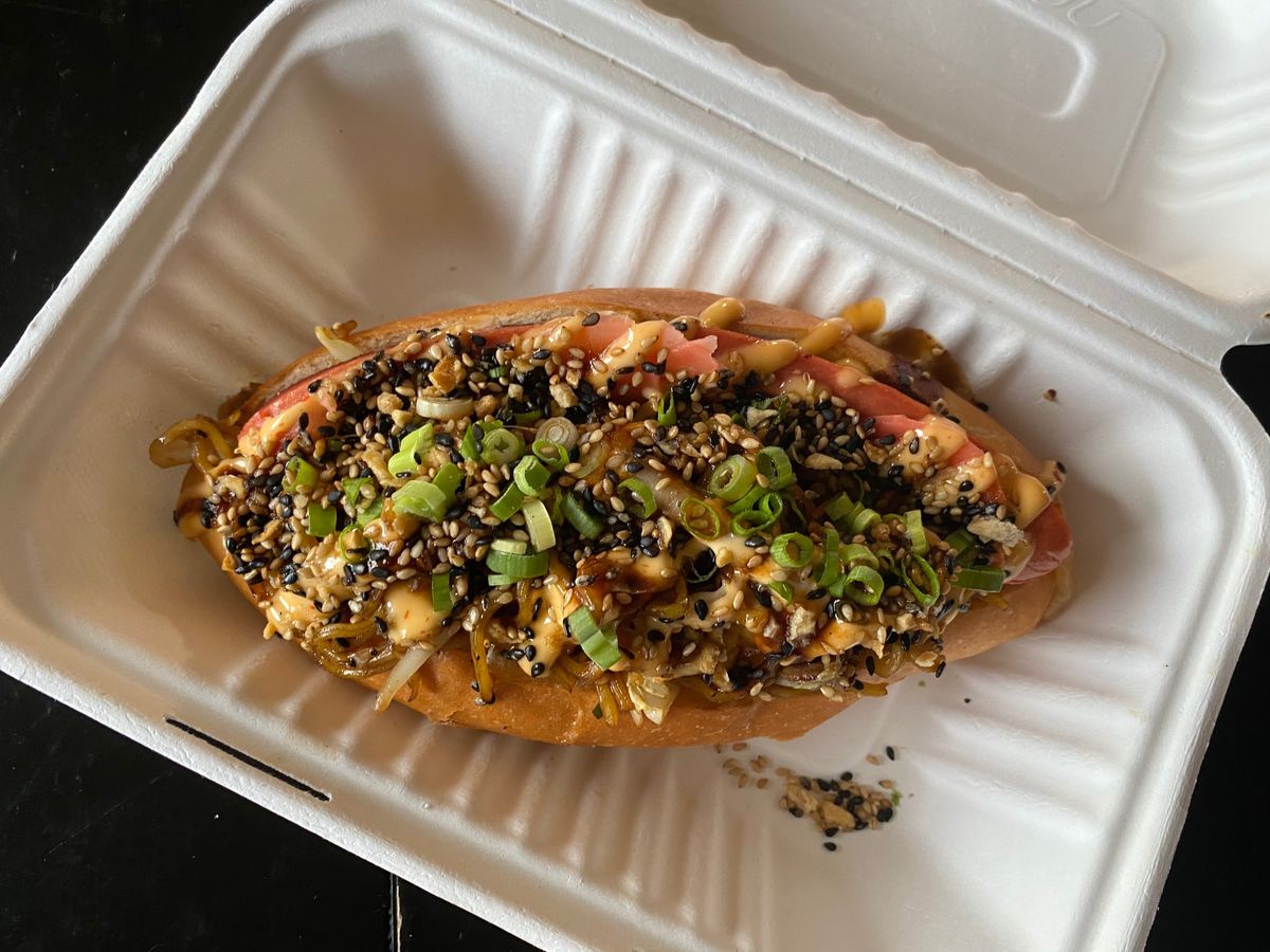 A yakisoba pan in a takeout box: a fat torpedo roll overstuffed with stir-fried noodles and garnished with scallions and sesame seeds