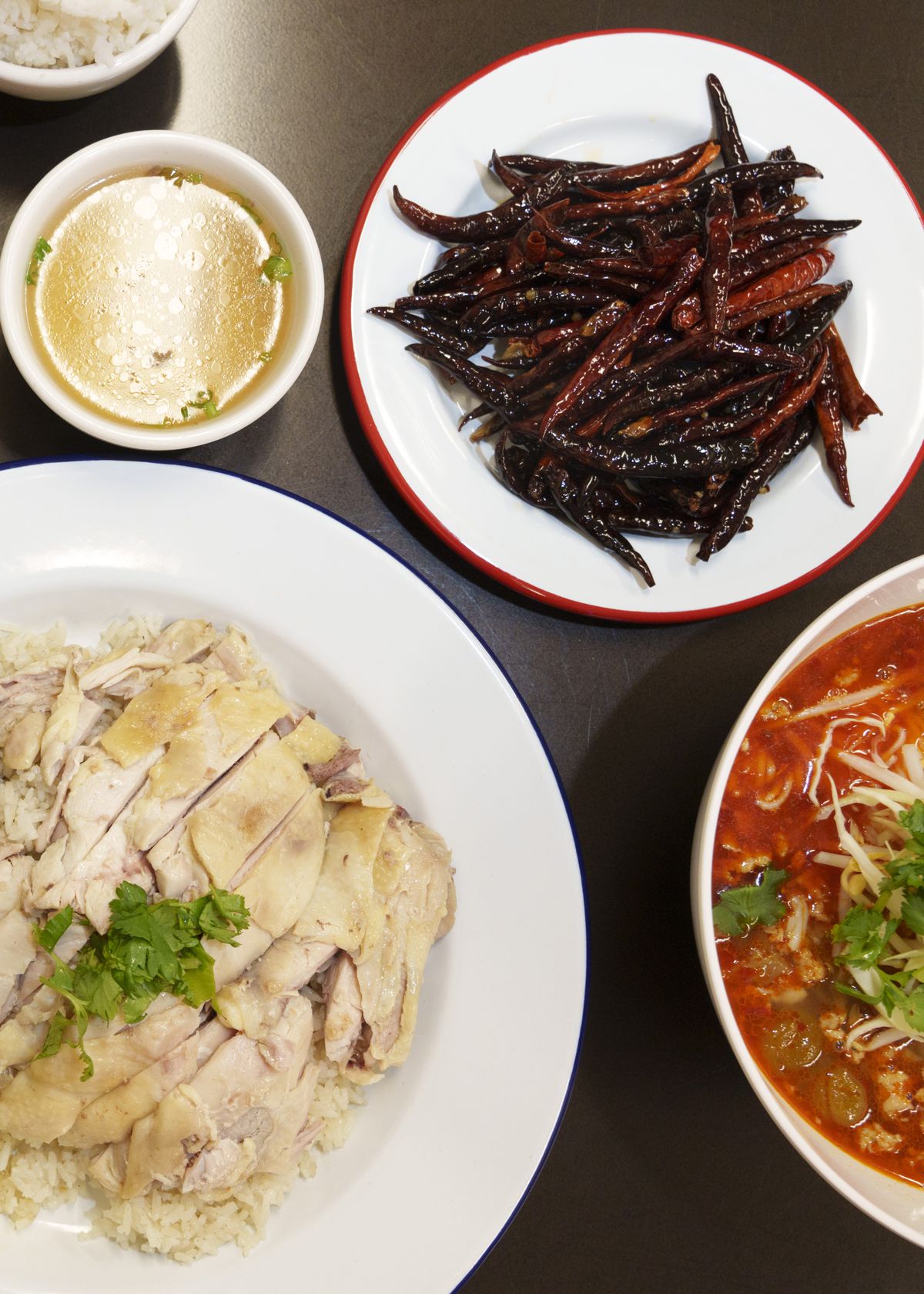 An aerial view of few white dishes on a black table: one of chicken and rice, one of dark red chiles, one a small cup of broth, one a bowl of bright red noodle soup. 