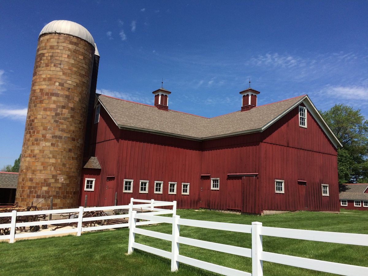 A traditional red barn with an attached grain silo surrounded by a grassy lawn and a white fence.