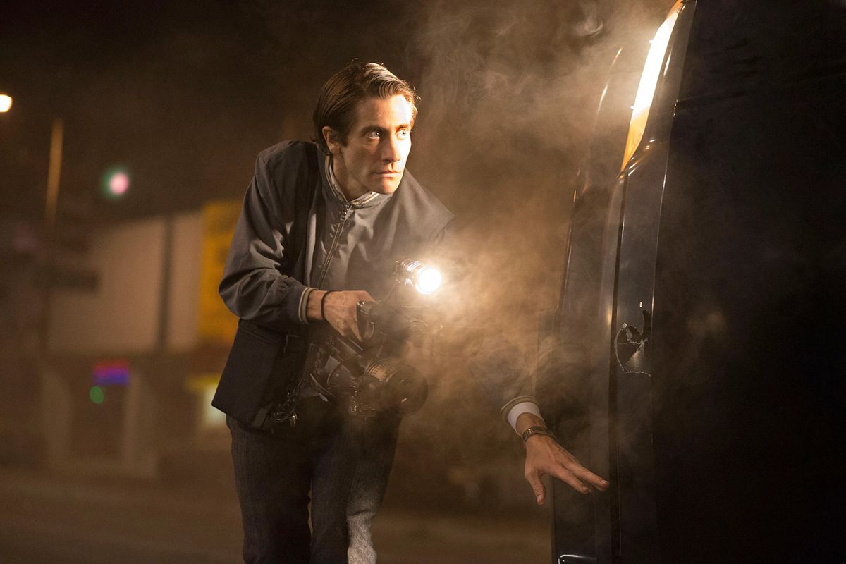 Jake Gyllenhaal as Louis “Lou” Bloom holding a camera behind an overturned car in Nightcrawler.