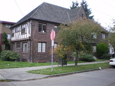 A brick building with a hipped roof has a brick front facade with a single, large gable in the center. On the left side, the first story is brick and the second is half-timbered. The entrance is hidden behind a medium-sized tree.